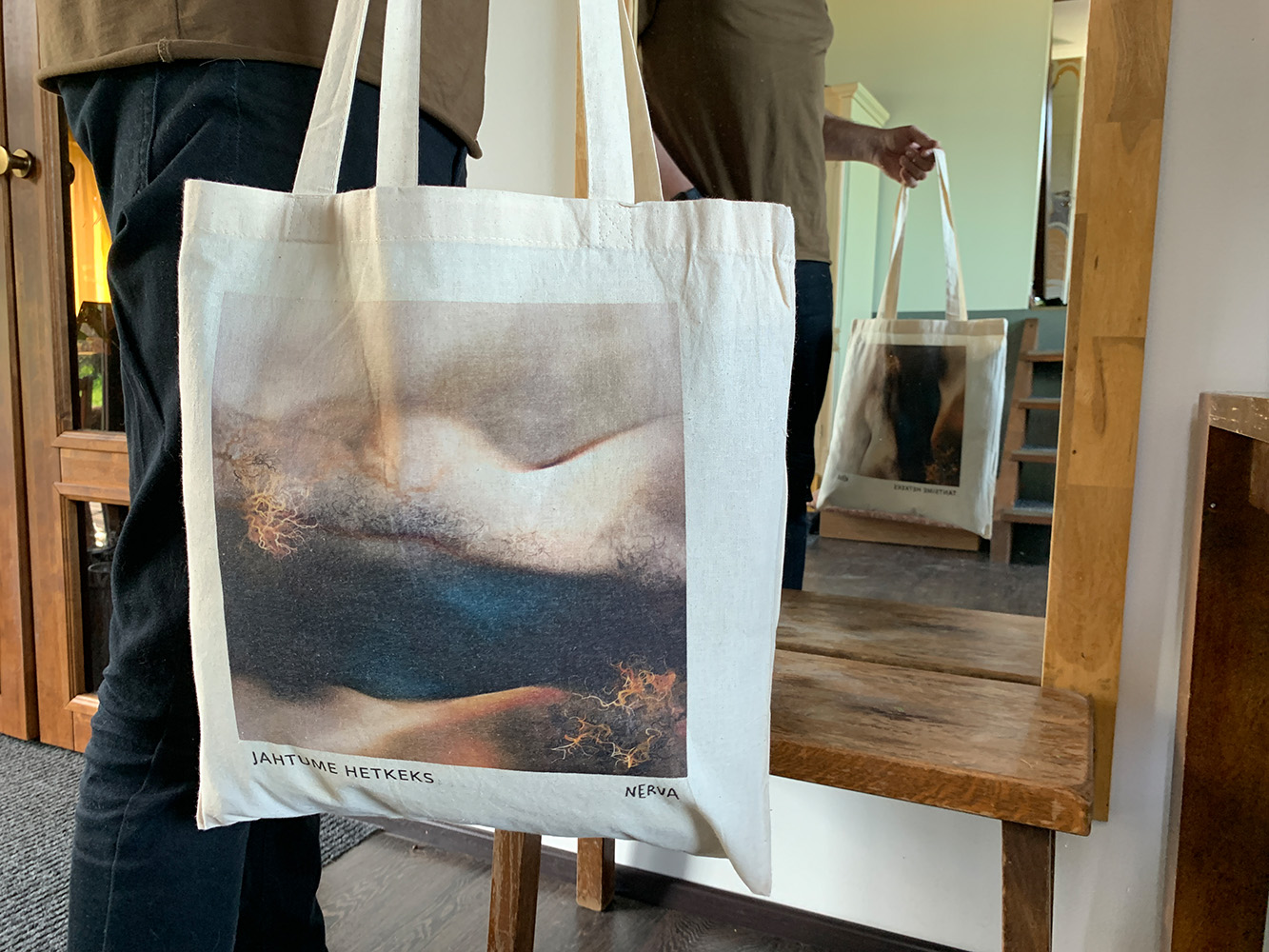 Experience the art of Nerva firsthand with this unique photo of the artist holding the 100% cotton campaign bag. This image captures a clever view in which both sides of the bag are visible, with 'Jahtume hetkeks' facing forward and 'Tantsime hetkeks' reflected in a mirror. The natural lighting and setting emphasize the bag’s quality and the vibrant, lasting print of the artwork.