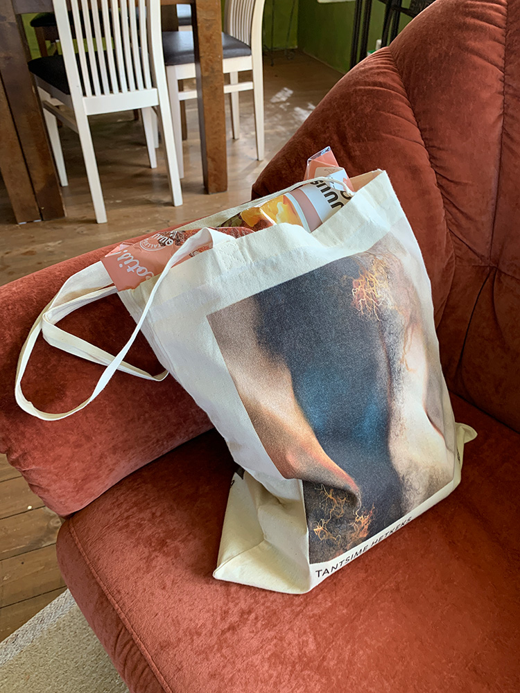 Captured just after a grocery run, this photo features the 'Tantsime hetkeks' side of the 100% cotton campaign bag, resting on a soft, rusty-red armchair near the dining room. The bag, filled to the brim with various items, prominently displays a package of three-cheese oven chicken, hinting at the day’s culinary adventures. The vibrant artwork on the bag contrasts beautifully with the chair’s warm tones, offering a glimpse into a day filled with art and indulgence.